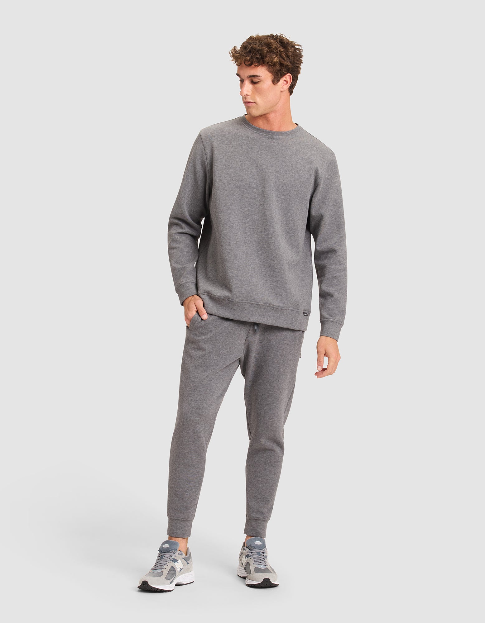 A man stands against a plain light background, looking left with one hand in his pocket, wearing Cozy Earth Men's StretchTech Joggers paired with a matching gray sweatshirt and gray and white sneakers. 