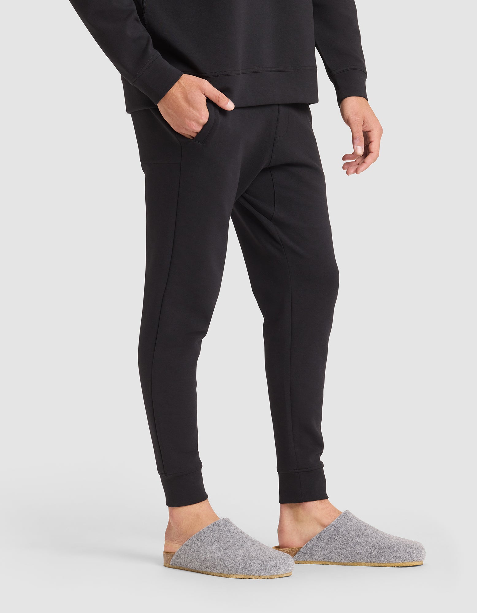 A person wearing Cozy Earth Men's StretchTech Jogger pants and a matching long-sleeve top stands with one hand in their pocket, wearing gray felt slippers with tan soles, on a white background. 