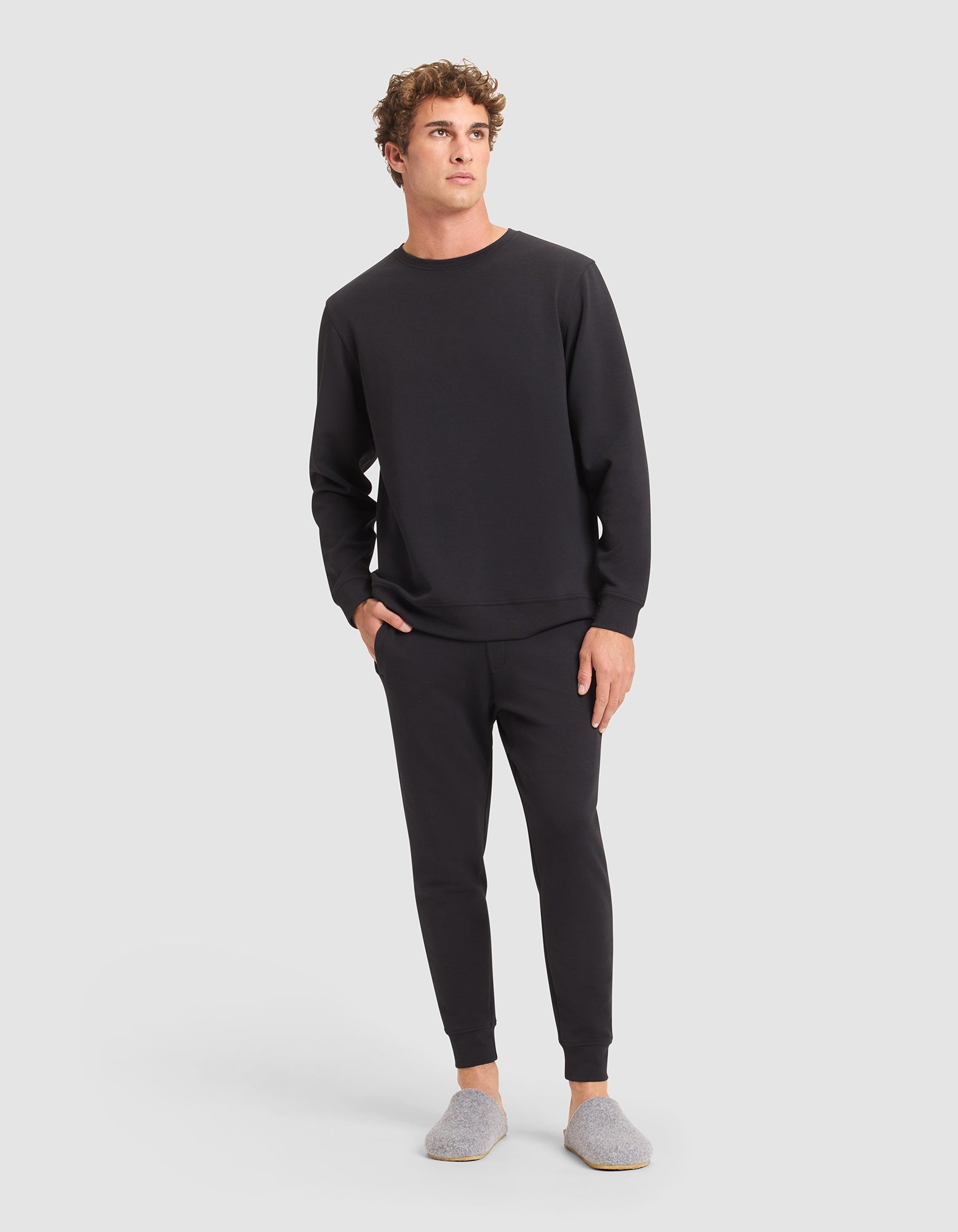 A man stands against a plain background wearing Cozy Earth’s Men's StretchTech Jogger in black, paired with a black sweatshirt and gray slippers. He has one hand in his pocket and looks slightly to the side. 
