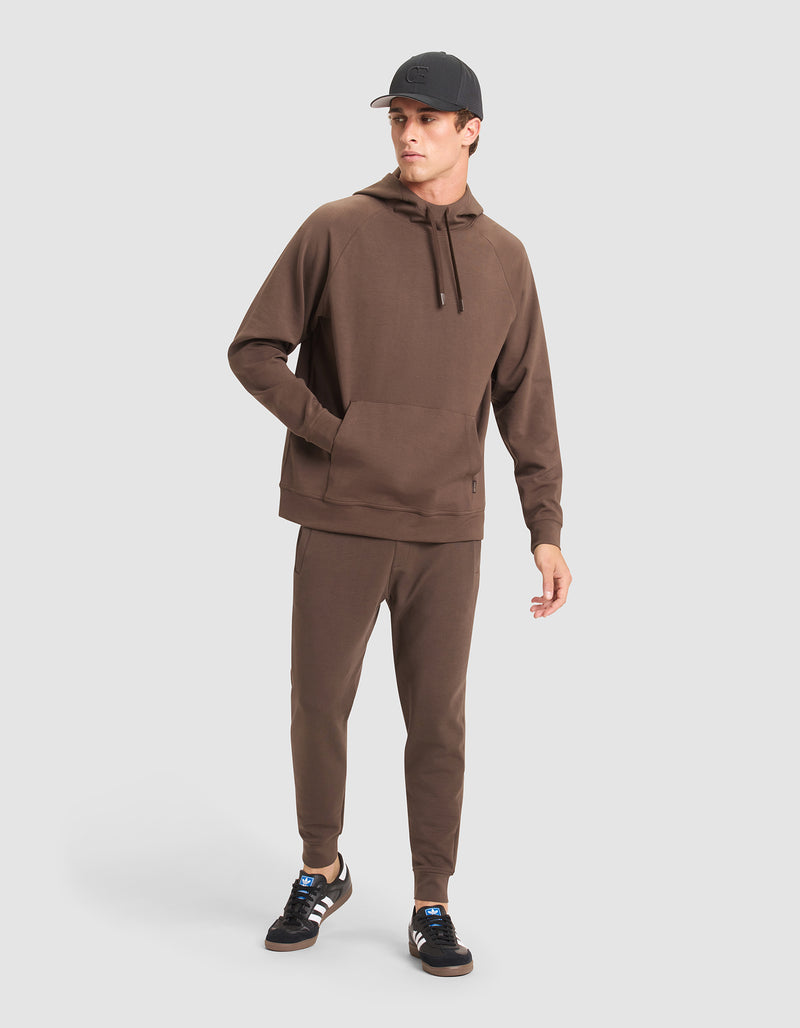 A man wears the Cozy Earth Men's StretchTech Hoodie & Jogger Set with a black cap and black Adidas sneakers featuring white stripes, standing against a plain light gray background.