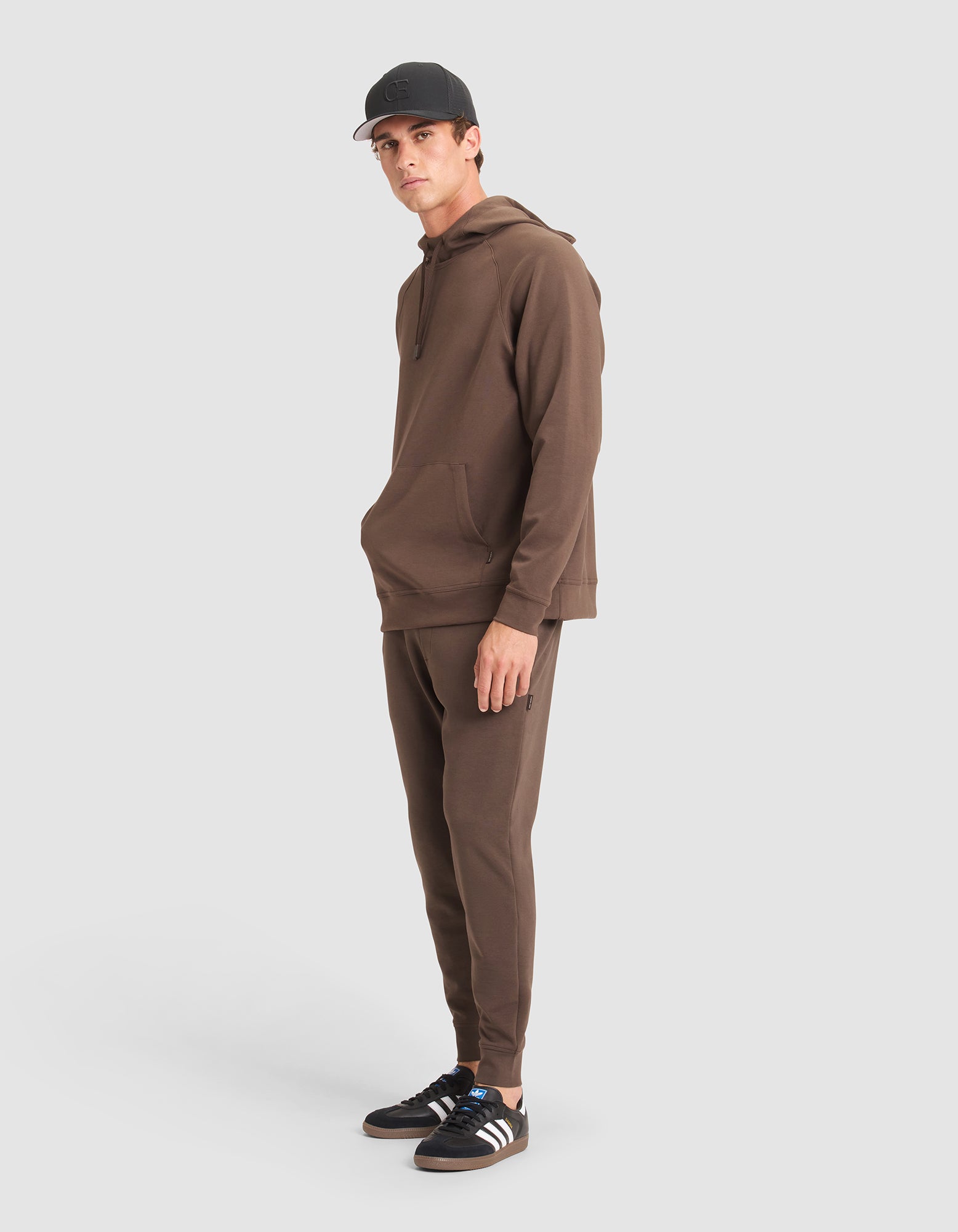 A man models the Cozy Earth Men's StretchTech Hoodie & Jogger Set in brown against a plain white background, paired with a black cap and black Adidas sneakers with white stripes. He stands with hands by his sides, gazing slightly to the side.  