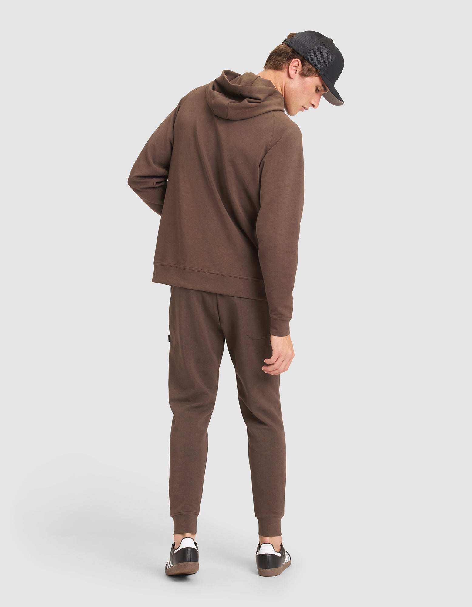 A man models the Cozy Earth Men's StretchTech Hoodie & Jogger Set in brown, paired with a black cap and black-and-white sneakers, standing against a plain light background and glancing over his shoulder.  |Color:Cacao