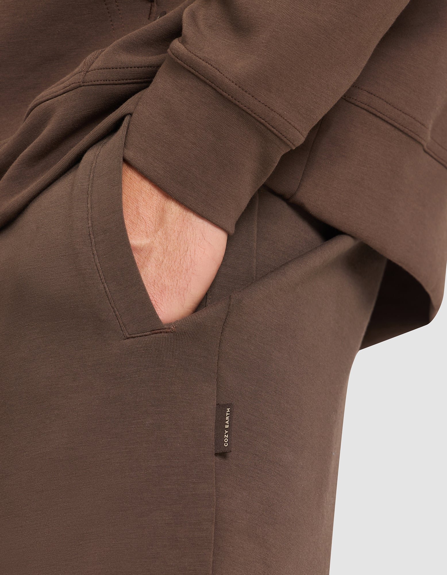 A person wears the Cozy Earth Men's StretchTech Hoodie & Jogger Set in brown, with one hand relaxed in the jogger pocket; a small tag on the pocket displays “COZY EARTH”.  