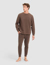 A young man with curly hair stands barefoot against a light gray backdrop, wearing the Cozy Earth Men's StretchTech Crewneck & Jogger Set in brown. |Color:Cacao