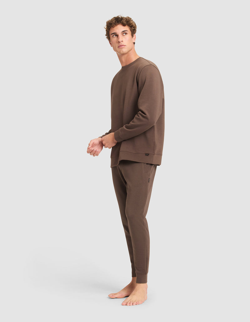 A person stands barefoot on a plain background, wearing the Cozy Earth Men's StretchTech Crewneck & Jogger Set in brown, looking slightly upward and to the side.