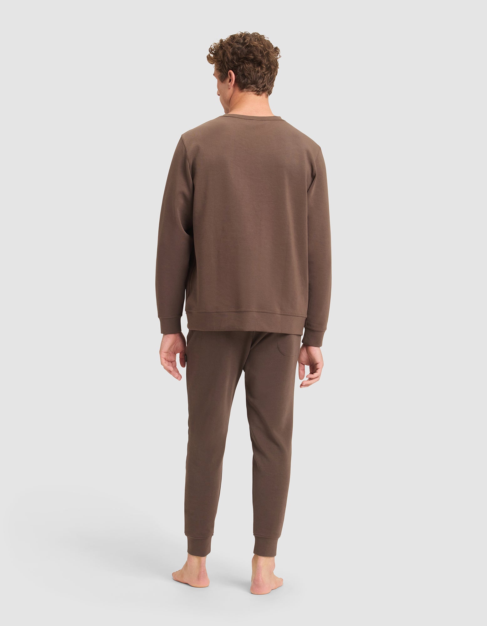 A man stands barefoot facing away, wearing the Cozy Earth Men's StretchTech Crewneck & Jogger Set in brown, set against a plain light gray background. |Color:Cacao