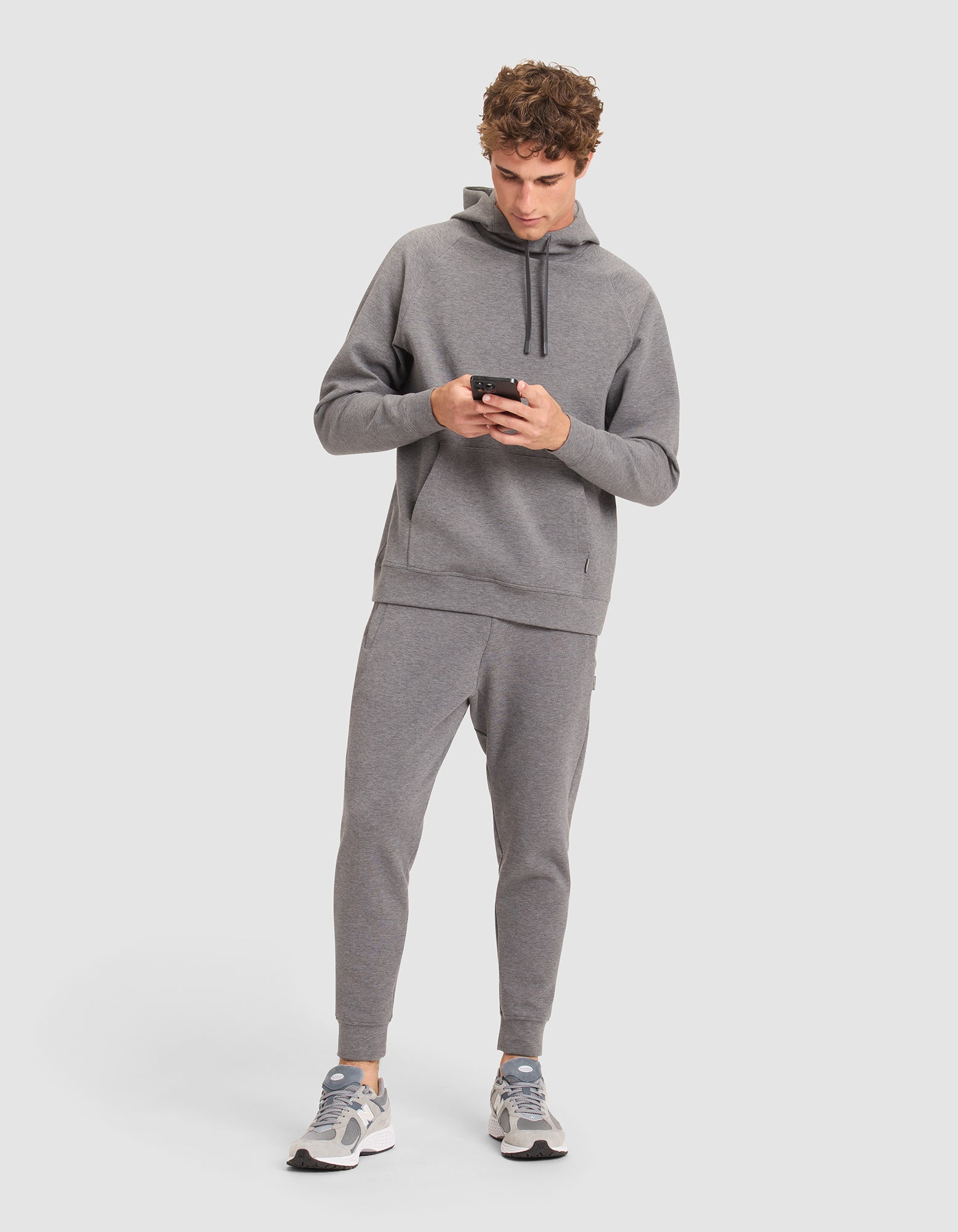 A man stands against a plain light background, looking at his phone while wearing Cozy Earth's Men's StretchTech Crewneck & Jogger Set in grey, paired with white and grey sneakers. 