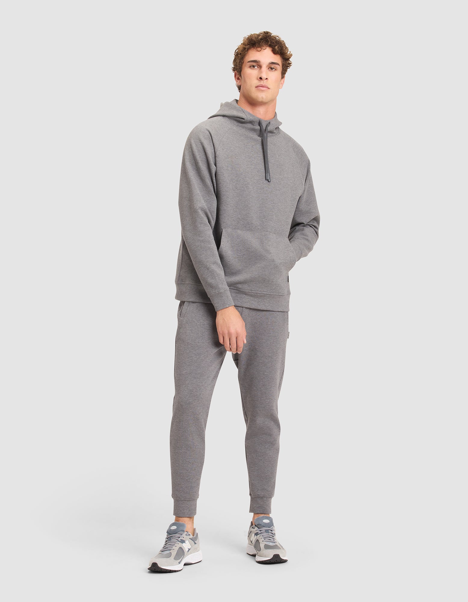 A young man faces forward wearing the Cozy Earth Men's StretchTech Hoodie & Jogger Set with light gray sneakers. His right hand is in his pocket, and he looks slightly upward against a plain light gray background. 