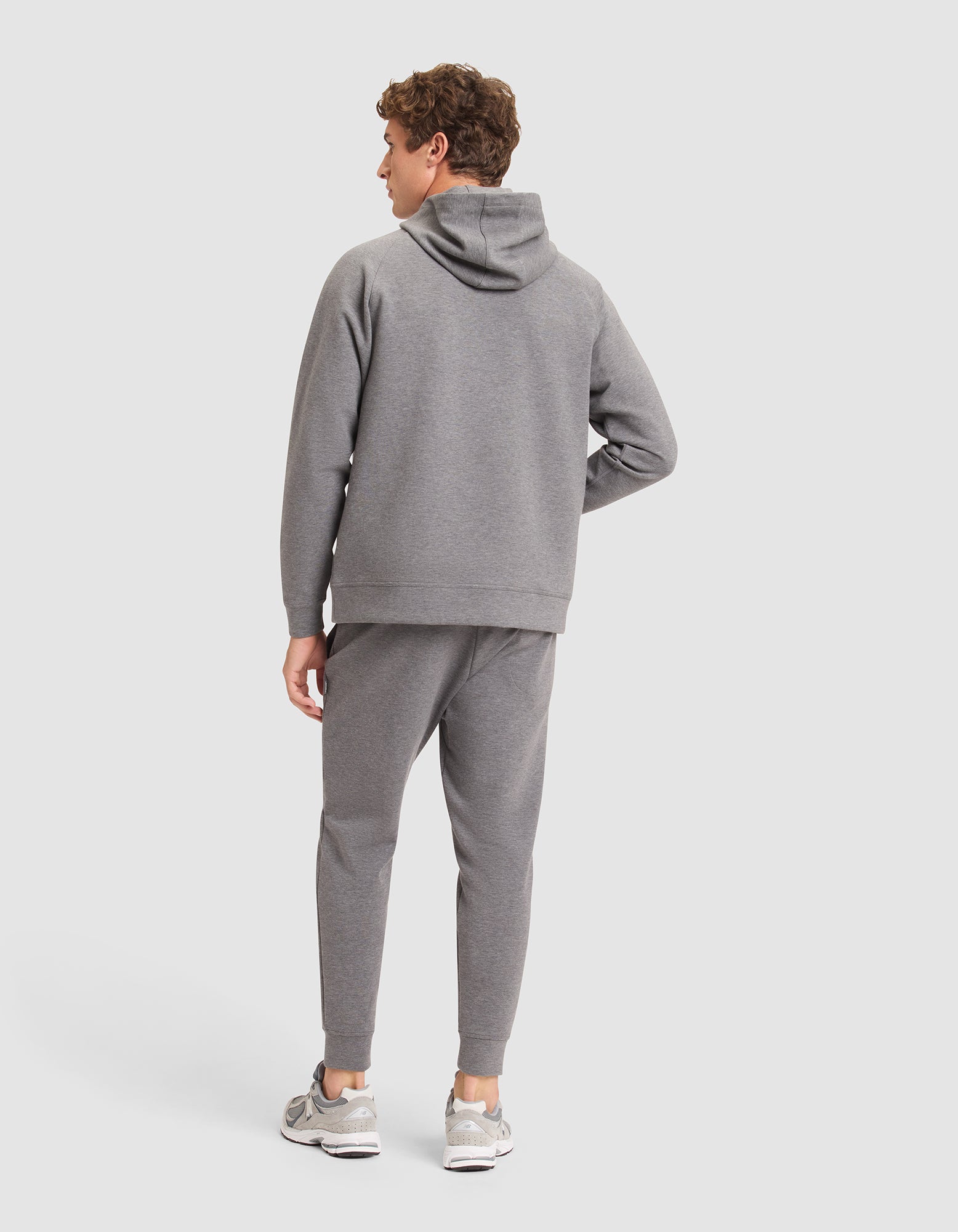 A man faces away from the camera, dressed in Cozy Earth's Men's StretchTech Hoodie & Jogger Set in gray, paired with gray sneakers against a plain light background. 