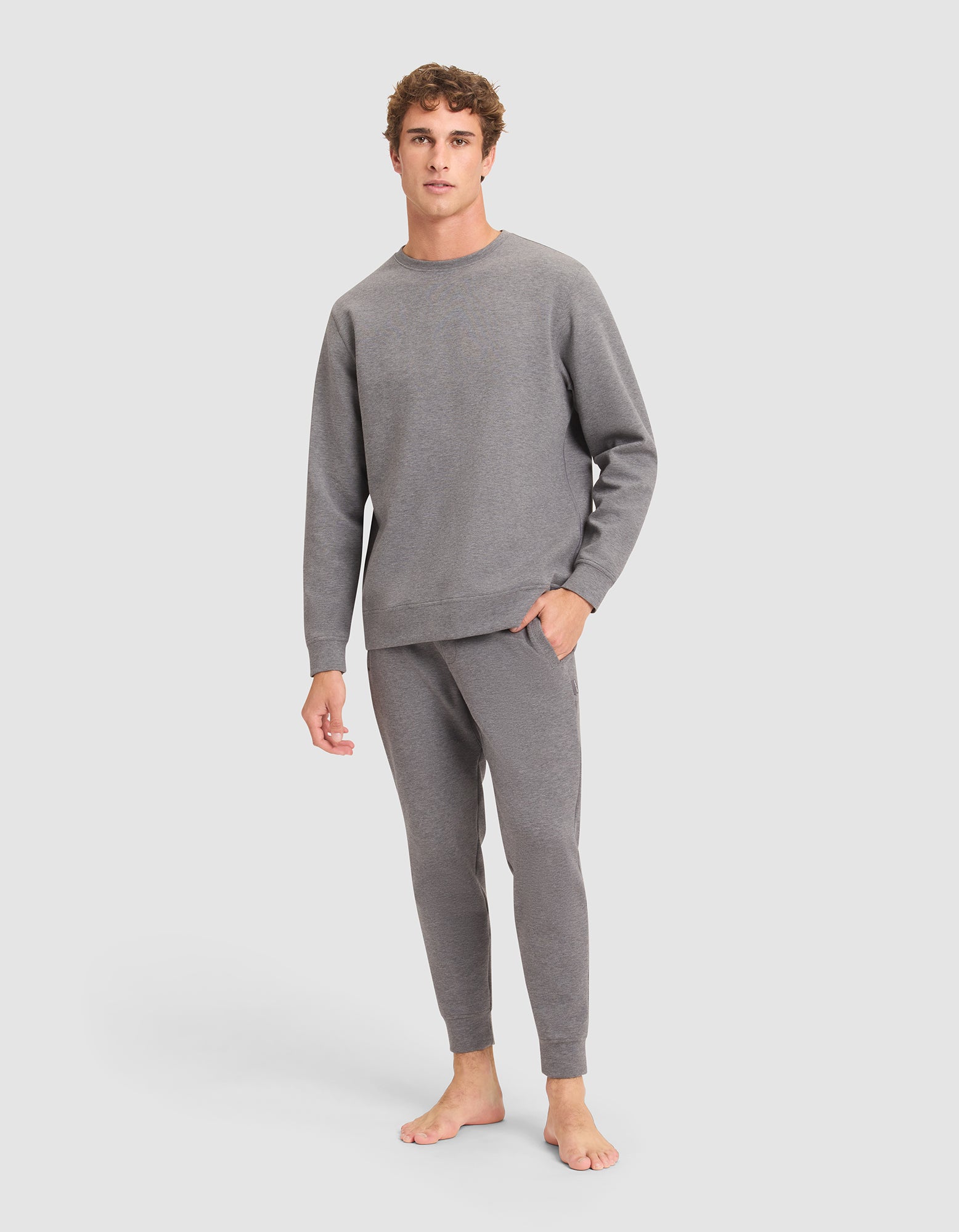 A young man with curly hair stands barefoot against a plain background, wearing the Cozy Earth Men's StretchTech Crewneck & Jogger Set in gray. 
