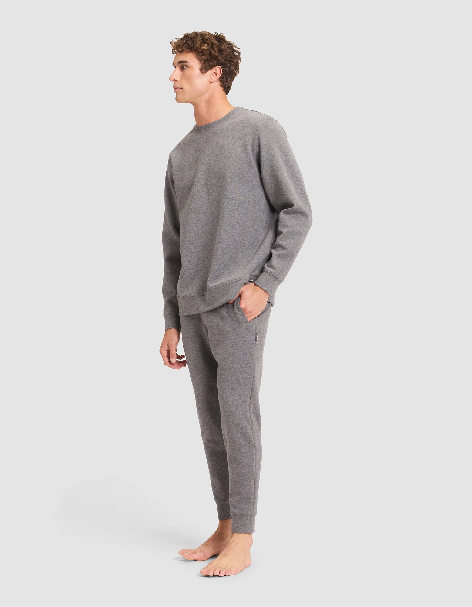 A person stands barefoot against a plain background, wearing the Cozy Earth Men's StretchTech Crewneck & Jogger Set in grey, with one hand in their pocket and looking to the side. 