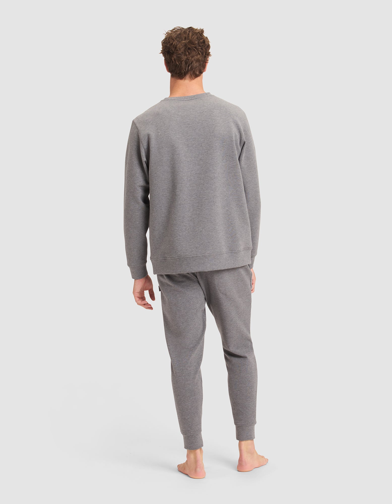A person stands barefoot facing away, wearing the Cozy Earth Men's StretchTech Crewneck & Jogger Set in light gray against a plain background. 