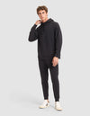 A young man with short curly hair poses against a light gray background, wearing the Cozy Earth Men's StretchTech Hoodie & Jogger Set in black with white sneakers, looking at the camera and touching his neck. |Color:Jet Black