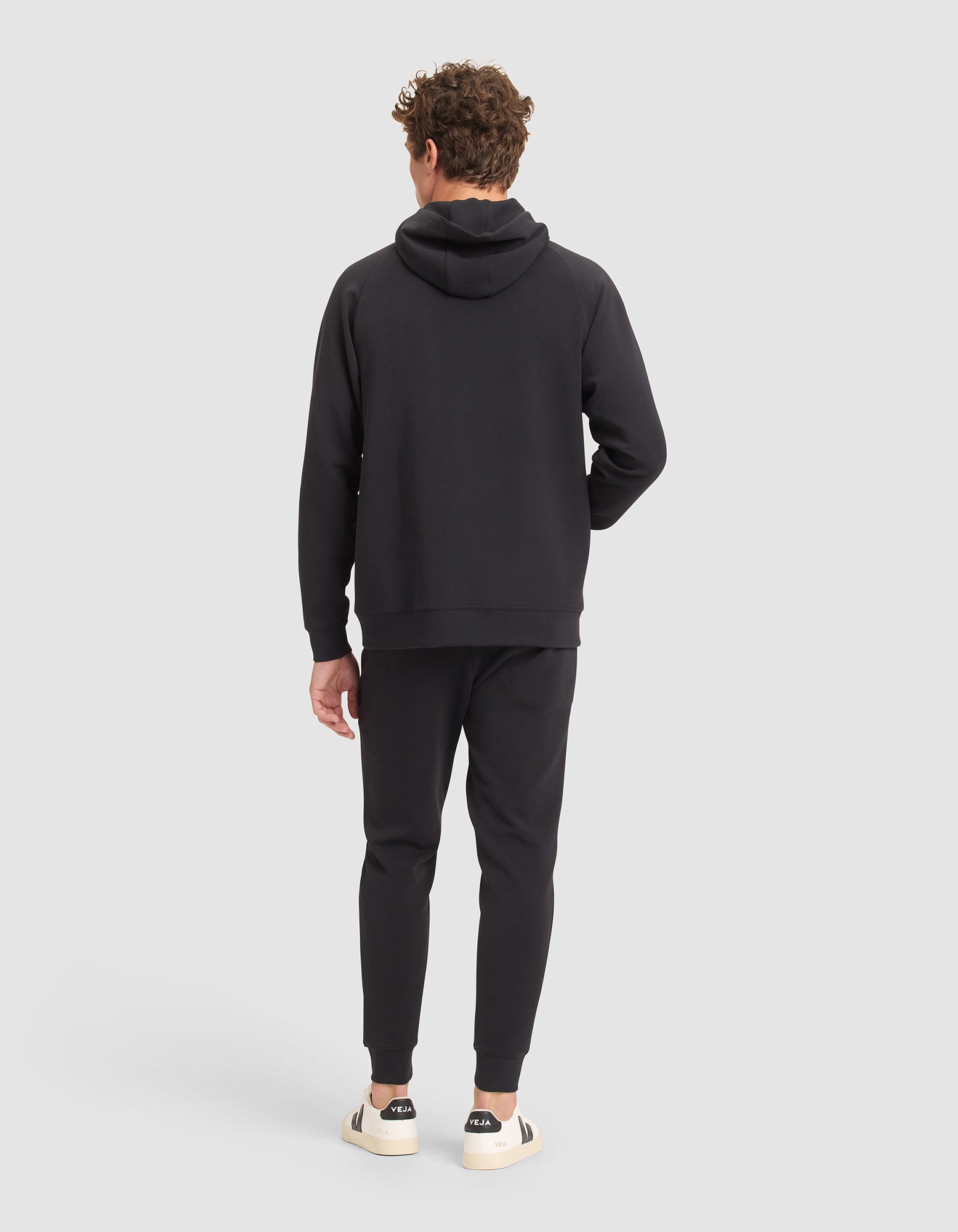 A man with curly hair faces away, wearing Cozy Earth's Men's StretchTech Hoodie & Jogger Set in black. He has one hand in his pocket and pairs the outfit with white sneakers featuring black accents against a plain light gray background. 