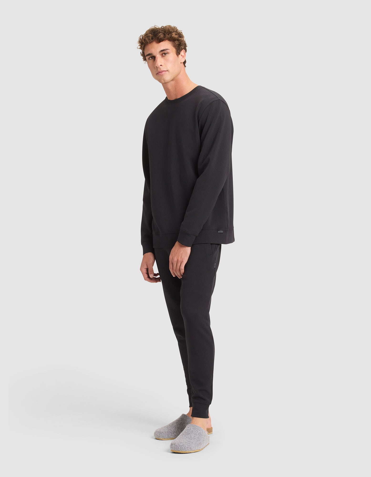 A young man models the Cozy Earth Men's StretchTech Crewneck & Jogger Set in black, paired with light gray slippers, standing against a plain light gray background and gazing slightly sideways at the camera. 