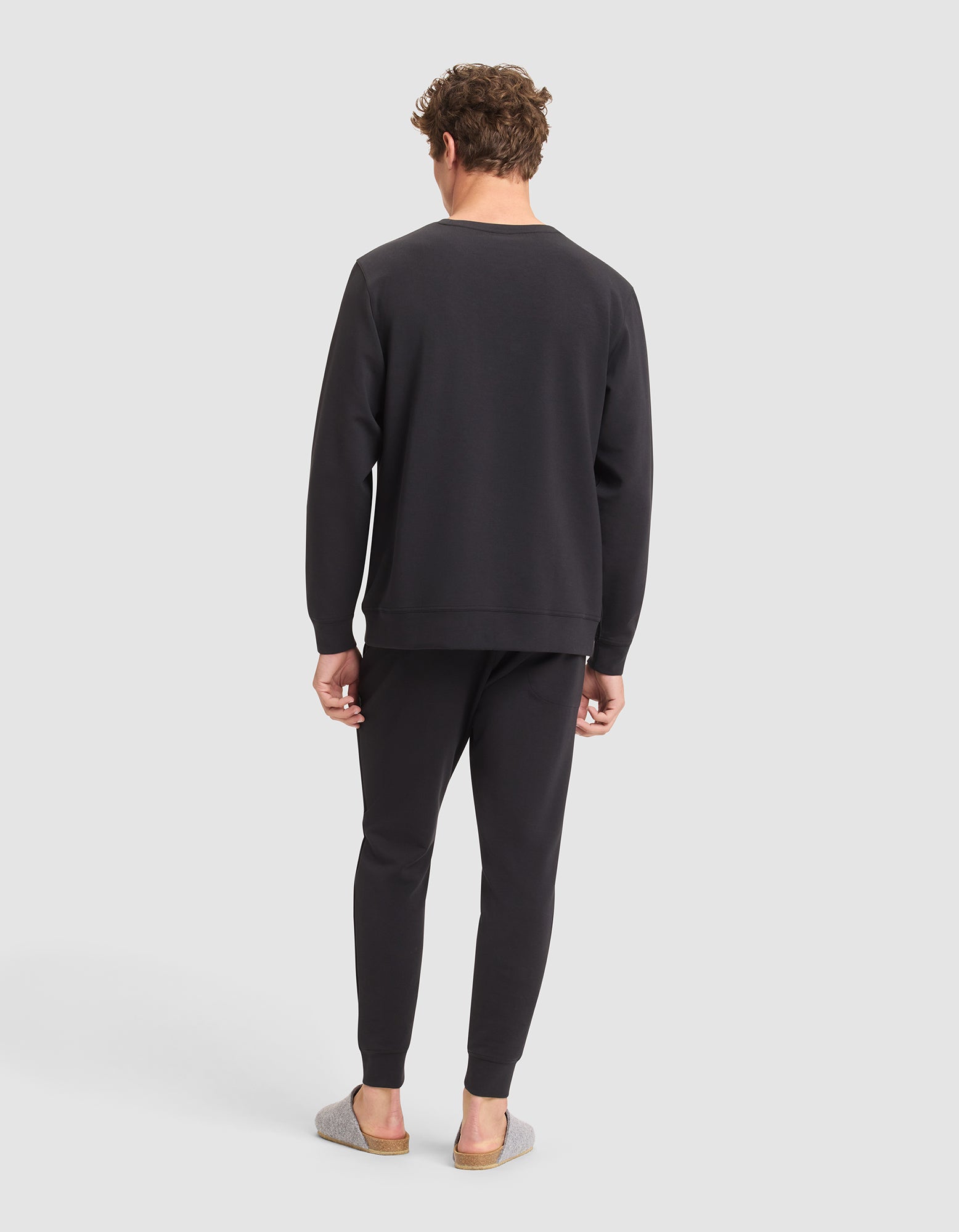 A man faces away from the camera, wearing the Cozy Earth Men's StretchTech Crewneck & Jogger Set in black with gray slip-on shoes, set against a plain light gray background. 