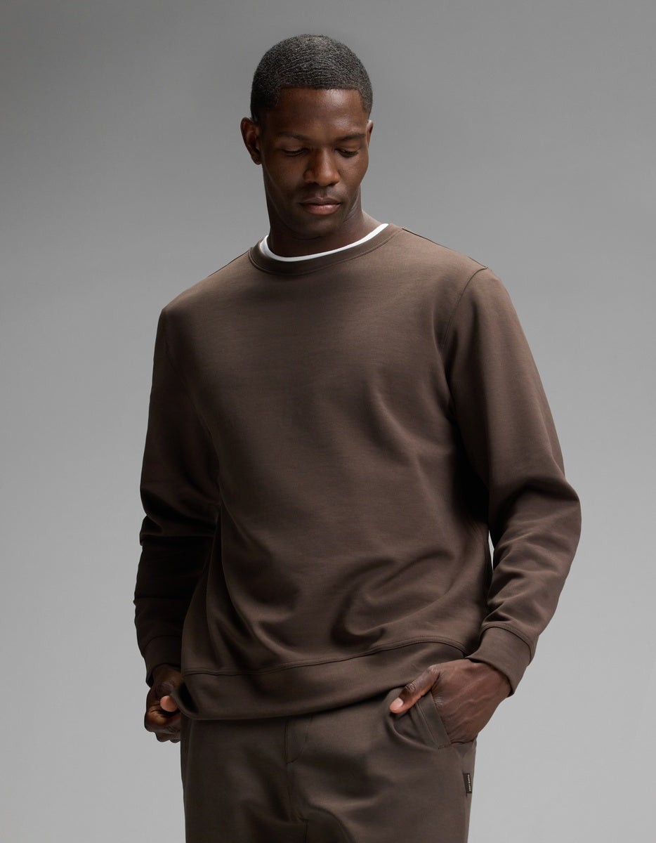 A man wearing the Cozy Earth Men’s StretchTech Crewneck in Cacao, paired with matching pants, stands with one hand in his pocket and a neutral expression against a plain gray background.|Color:Cacao