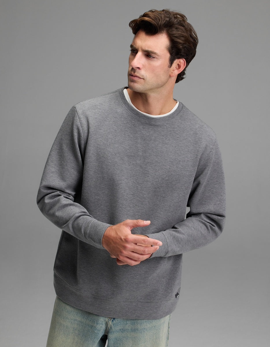 A man with short brown hair wears the Cozy Earth Men’s StretchTech Crewneck in Coal Heather and light blue jeans, standing against a plain gray background while looking to the side with his hands loosely clasped.|Color:Coal Heather