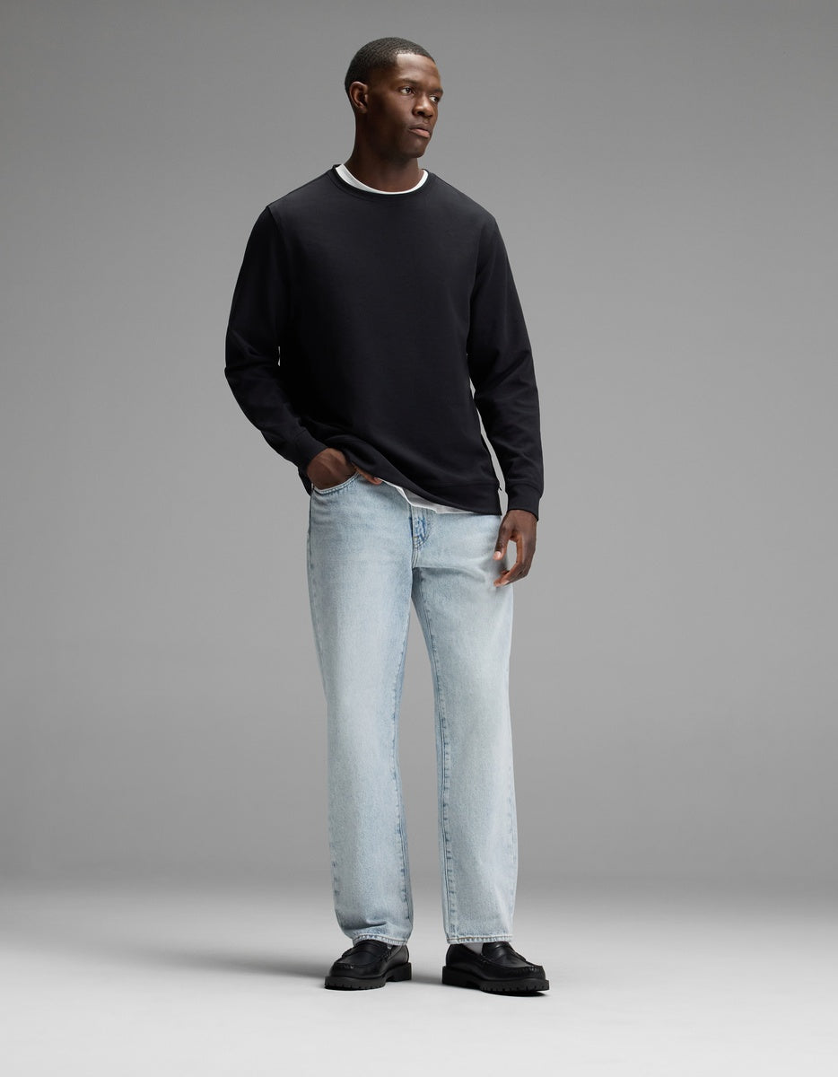 A man stands against a plain gray background, wearing the Cozy Earth Men’s StretchTech Crewneck in black over a white undershirt, paired with light blue jeans and black shoes. He has one hand in his pocket and looks slightly to his left.