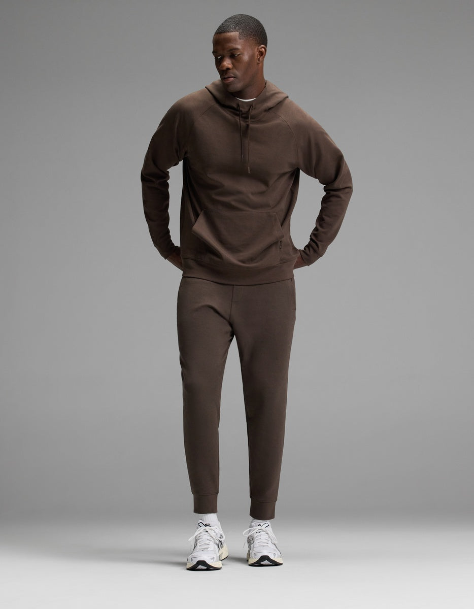 A man stands against a plain gray background, wearing the Cozy Earth Men’s StretchTech Hoodie & Jogger Set in brown with white sneakers, looking slightly down and to the side with his hands in his pockets.
