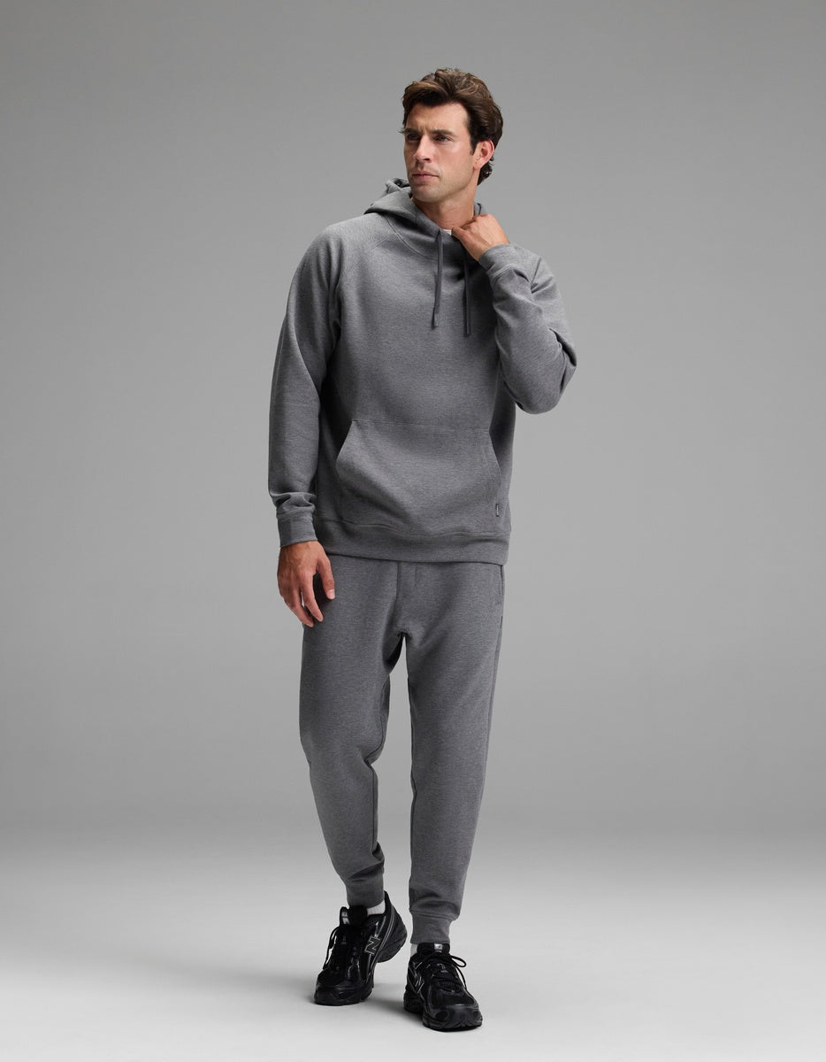 A man wearing the Cozy Earth Men’s StretchTech Hoodie & Jogger Set in gray stands against a plain gray background, looking to the side with one hand touching his hoodie, and wearing black sneakers.|Color:Coal Heather