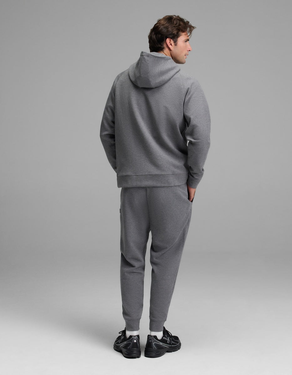 A person stands facing away, wearing the Cozy Earth Men’s StretchTech Hoodie & Jogger Set in gray with hands in pockets. The background is plain and neutral, and they are wearing black athletic sneakers.|Color:Coal Heather