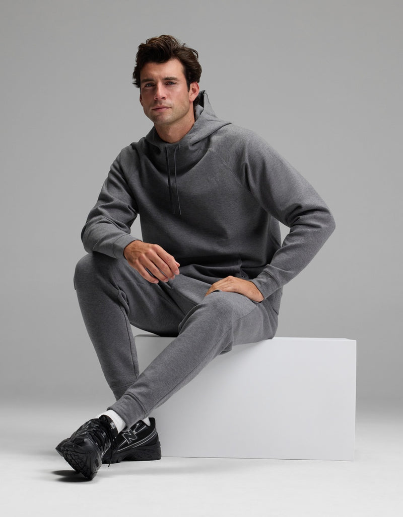 A man models the Cozy Earth Men’s StretchTech Hoodie & Jogger Set in gray, sitting on a white block against a plain gray background. He looks forward with a neutral expression and pairs the outfit with black sneakers.