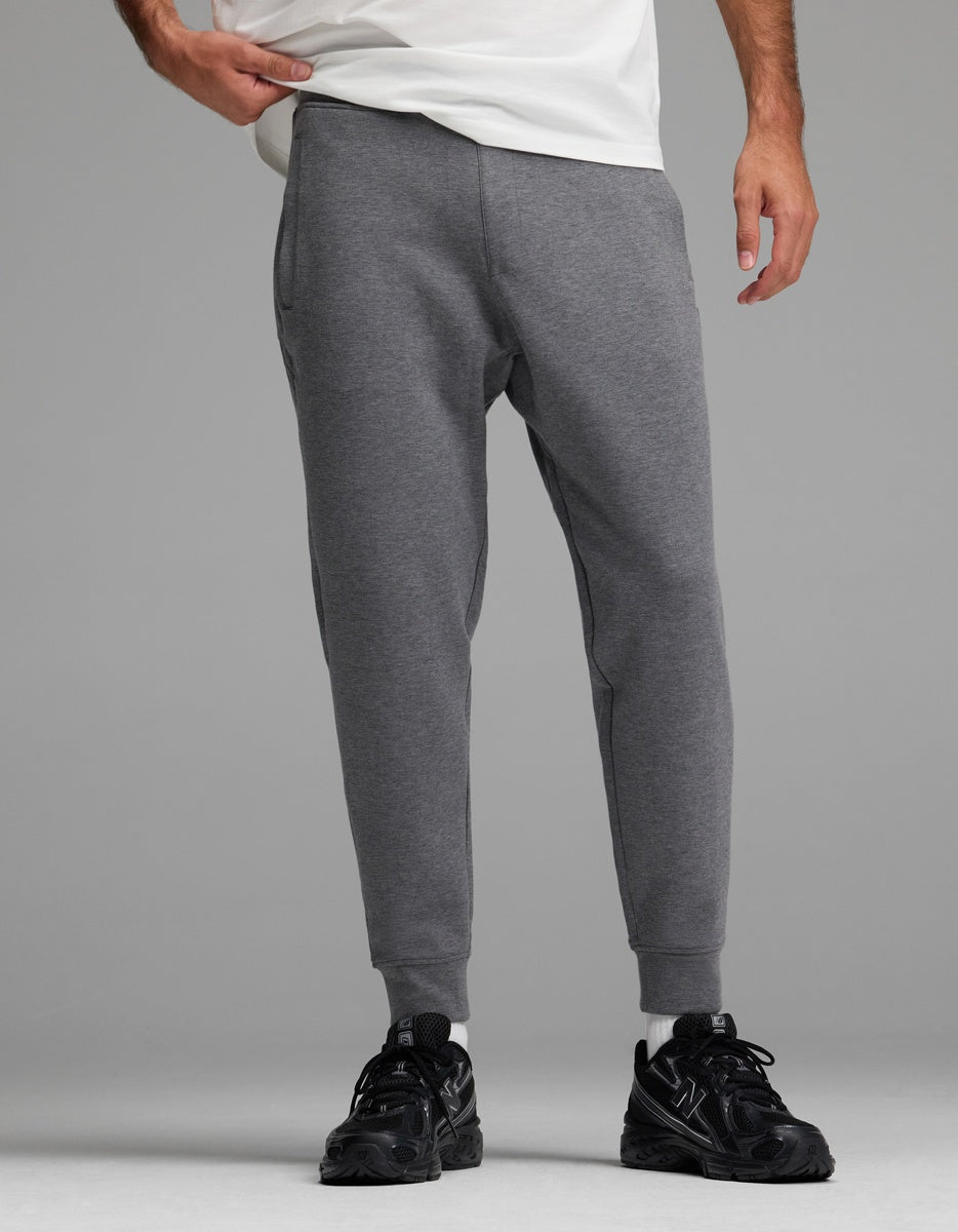 A person stands against a plain gray background, wearing Cozy Earth Men’s StretchTech Jogger sweatpants in gray, paired with black sneakers and a white shirt. Only the lower half of the body is visible.