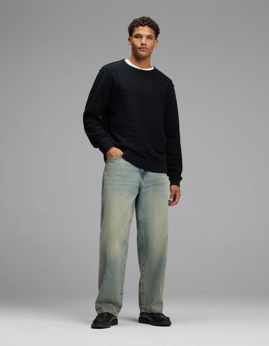 A man wearing a Cozy Earth Men’s Tate Crewneck over a white t-shirt, light-wash jeans, and black shoes.