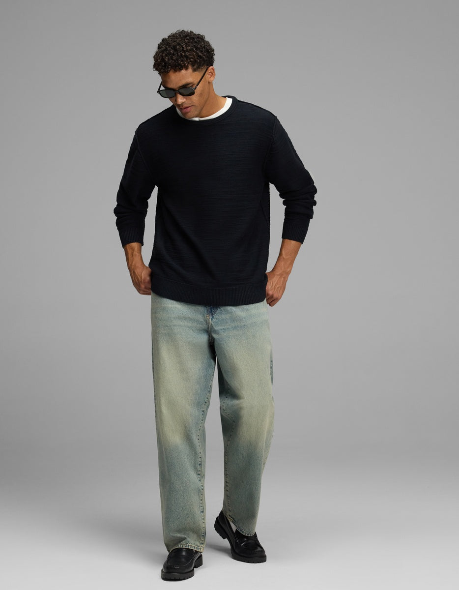 A person wearing a navy Cozy Earth Men’s Tate Crewneck, light-wash loose-fit jeans, and black loafers.