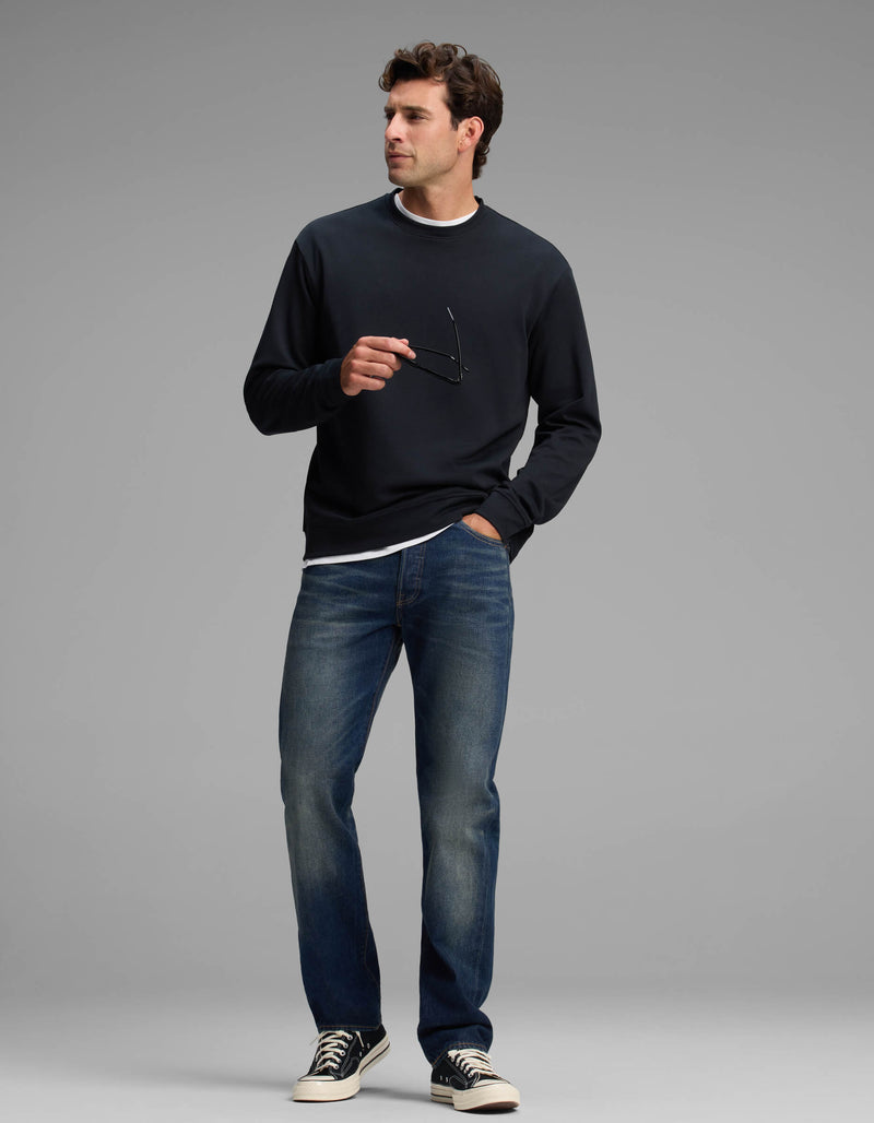A man wearing the Cozy Earth Men's Versa Crewneck, paired with blue jeans and black sneakers, stands holding eyeglasses in one hand while looking to the side against a plain gray background.
