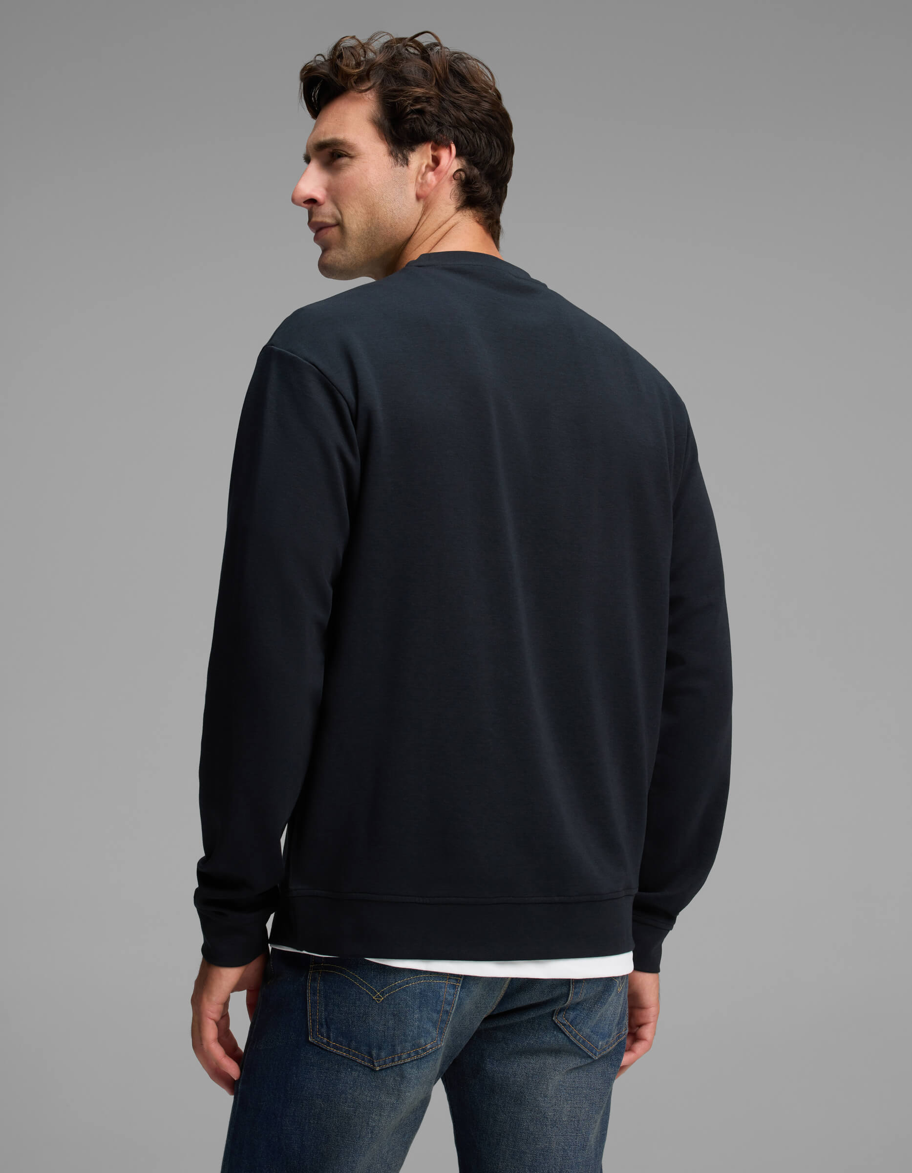A man with dark hair models the Cozy Earth Men's Versa Crewneck in black with blue jeans, standing with his back to the camera and looking over his left shoulder against a plain gray background.|Color:Jet Black