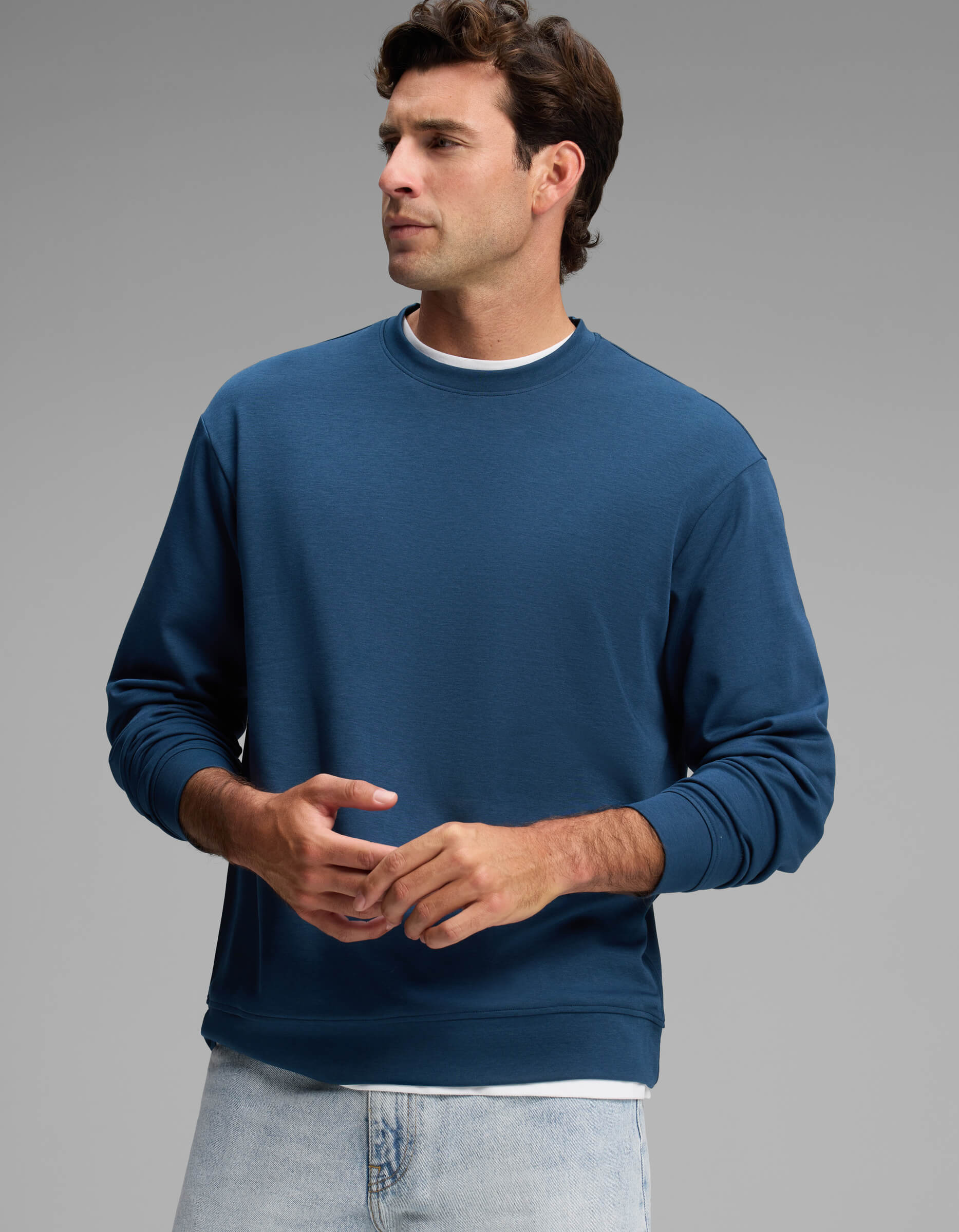 A man wearing the Cozy Earth Men's Versa Crewneck in blue over a white shirt and light blue jeans stands against a plain gray background, looking to his left with his hands loosely clasped.