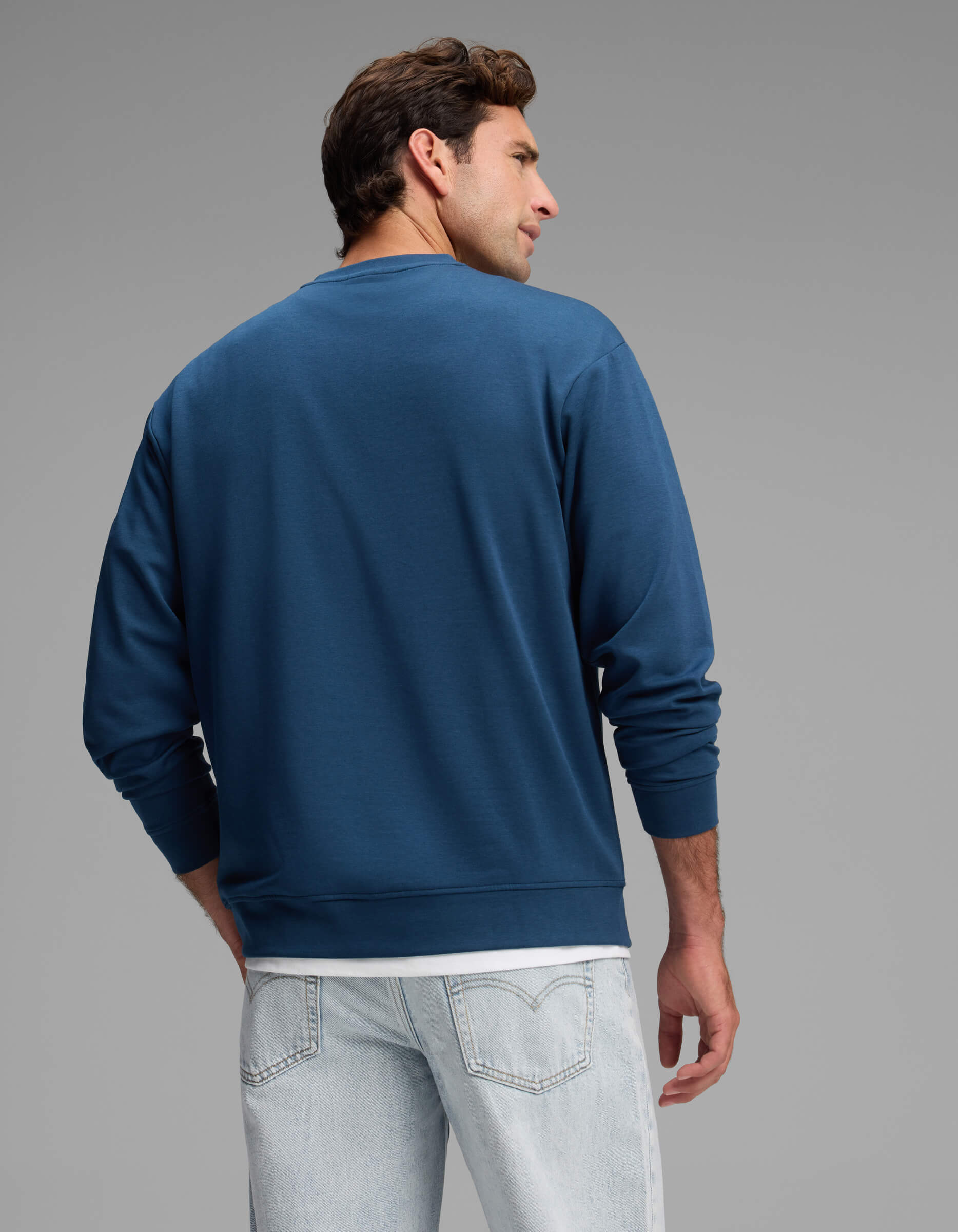 A man is shown from behind, wearing Cozy Earth’s Men's Versa Crewneck in blue over a white t-shirt and light-wash jeans, standing against a plain gray background.