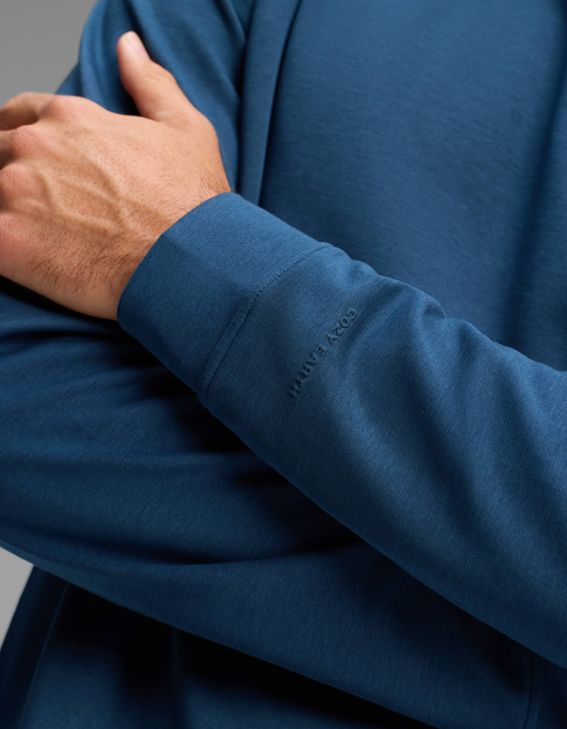 A person wearing the Men's Versa Crewneck by Cozy Earth in blue folds one arm across their chest. The shirt features a subtle "COZY EARTH" embossing near the wrist.