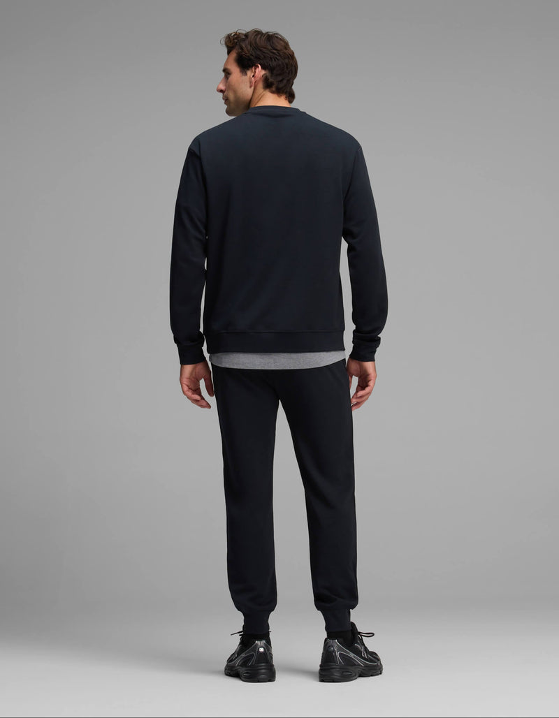A man is standing with his back to the camera, wearing the Cozy Earth Men's Versa Crewneck & Jogger Set in black, paired with black sneakers and a gray shirt peeking out underneath. The background is plain and light gray.