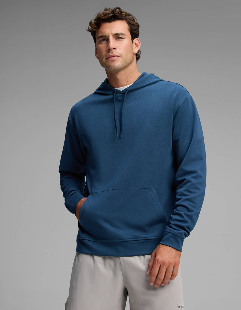 A man wearing the Cozy Earth Men's Versa Hoodie in blue and light gray shorts stands with one hand in his pocket against a plain gray background.