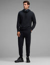 A man stands against a plain gray background, wearing the Cozy Earth Men's Versa Hoodie & Jogger Set in black, paired with black sneakers and over-ear headphones around his neck, looking slightly to the side.|Color:Jet Black