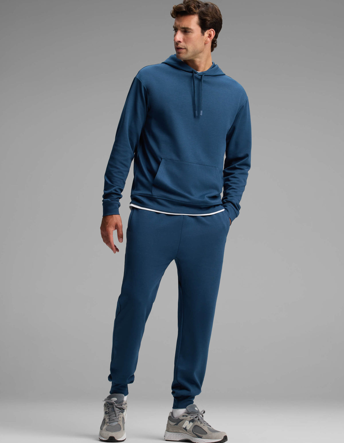 A man dressed in the Cozy Earth Men's Versa Hoodie & Jogger Set stands with one hand in his pocket, looking to the side. He completes the look with a white undershirt and gray sneakers.|Color:Obsidian