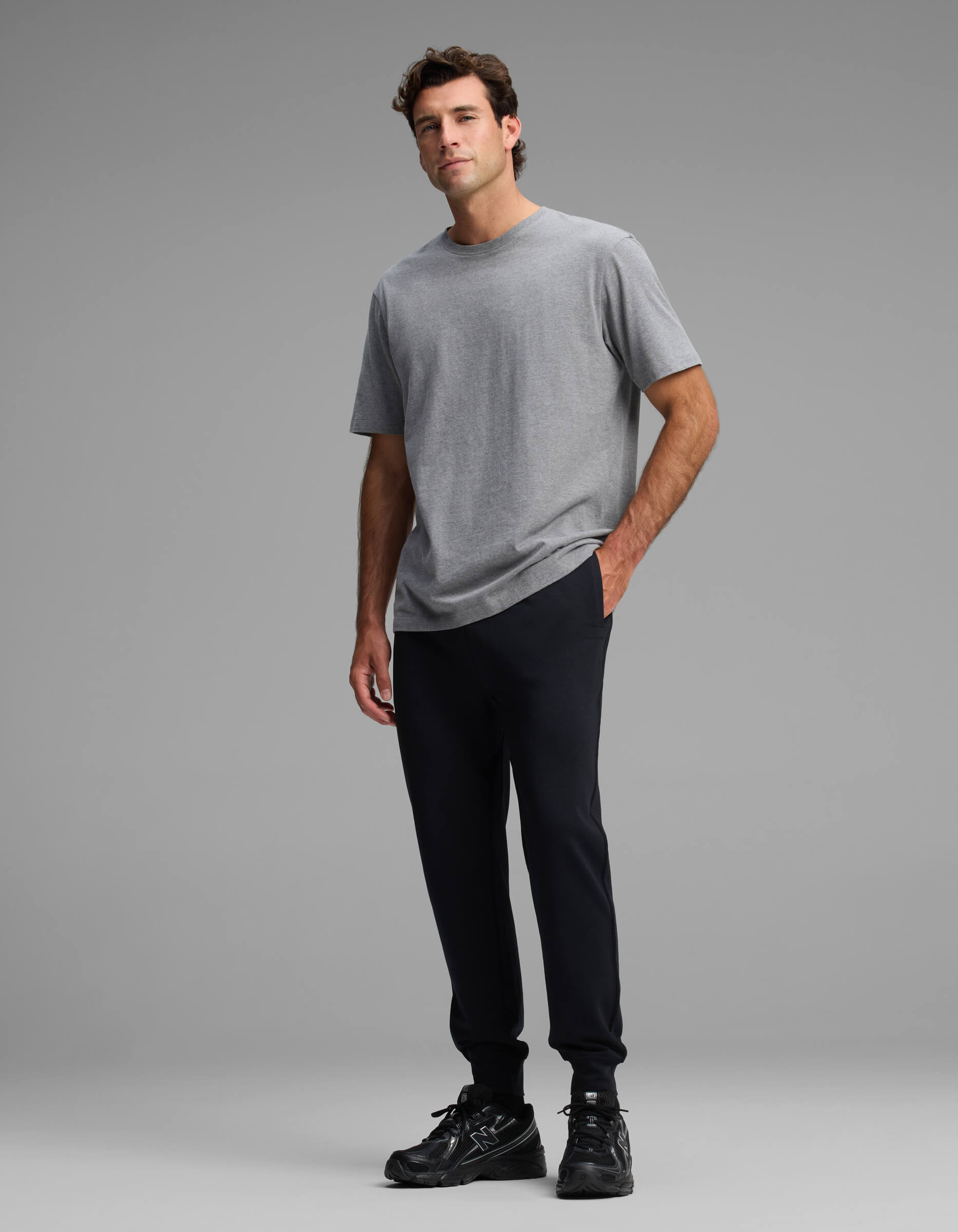 A man stands against a plain gray background, wearing a gray short-sleeve t-shirt, black sneakers, and the Cozy Earth Men's Versa Jogger in black, with one hand in his pocket.