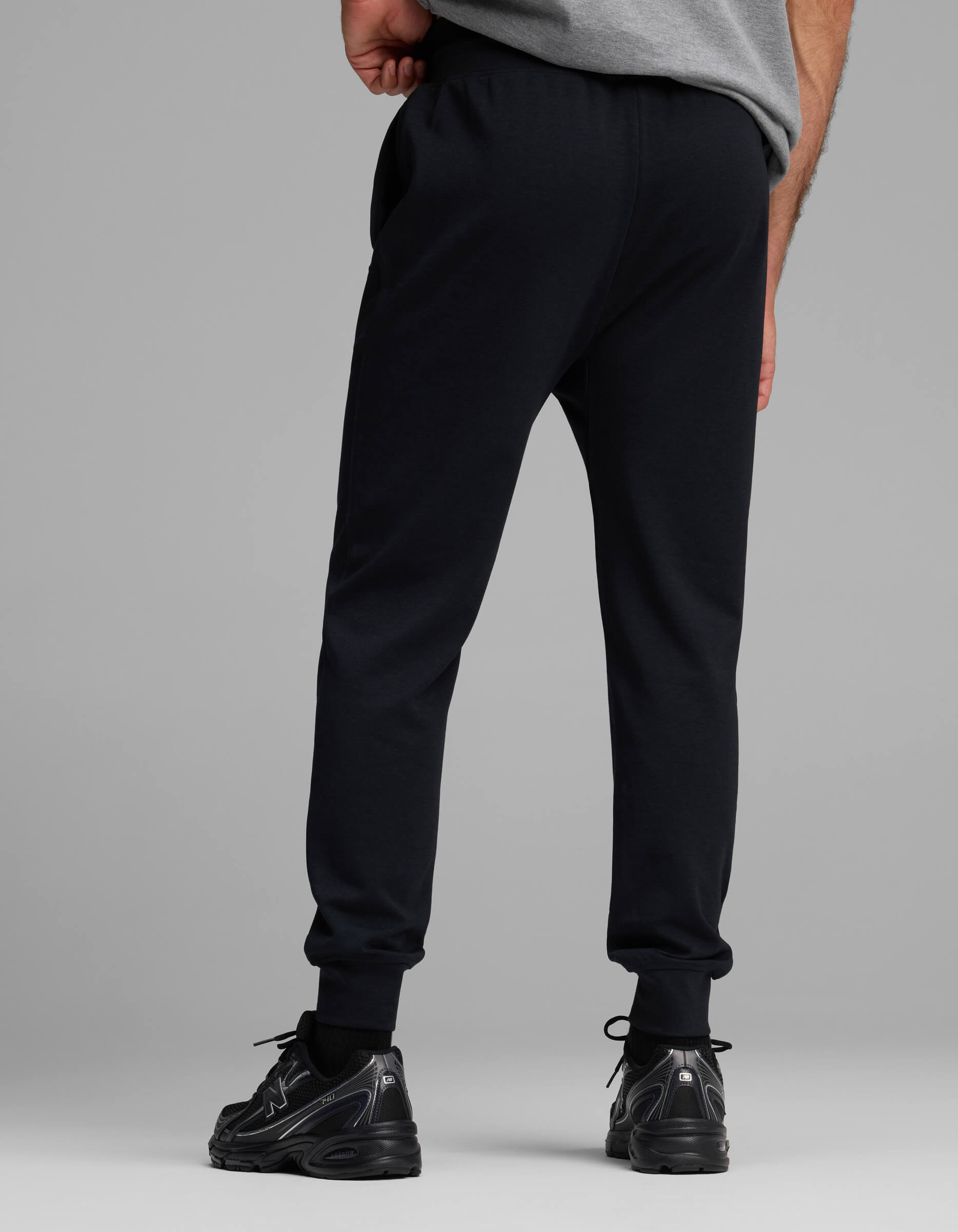 A person stands facing away, wearing a gray T-shirt, Cozy Earth Men's Versa Jogger sweatpants in black, and black sneakers against a plain gray background.