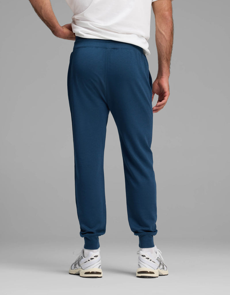 A person is standing with their back to the camera against a plain gray background, wearing a white T-shirt, white sneakers, and blue Cozy Earth Men's Versa Jogger pants.