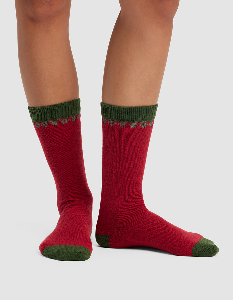 A pair of legs wear Cozy Earth's The Mistletoe Plush Lounge Socks, featuring red with green heels, toes, and cuffs plus a scalloped green and red design near the top. Set against a plain light gray background.