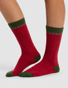 A person wears The Mistletoe Plush Lounge Sock by Cozy Earth, featuring red with green heels, toes, cuffs, and a green decorative top pattern, standing on a light gray background.|Color:Cranberry