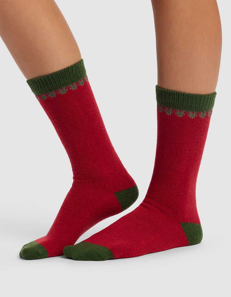 A person wears The Mistletoe Plush Lounge Sock by Cozy Earth, featuring red with green heels, toes, cuffs, and a green decorative top pattern, standing on a light gray background.