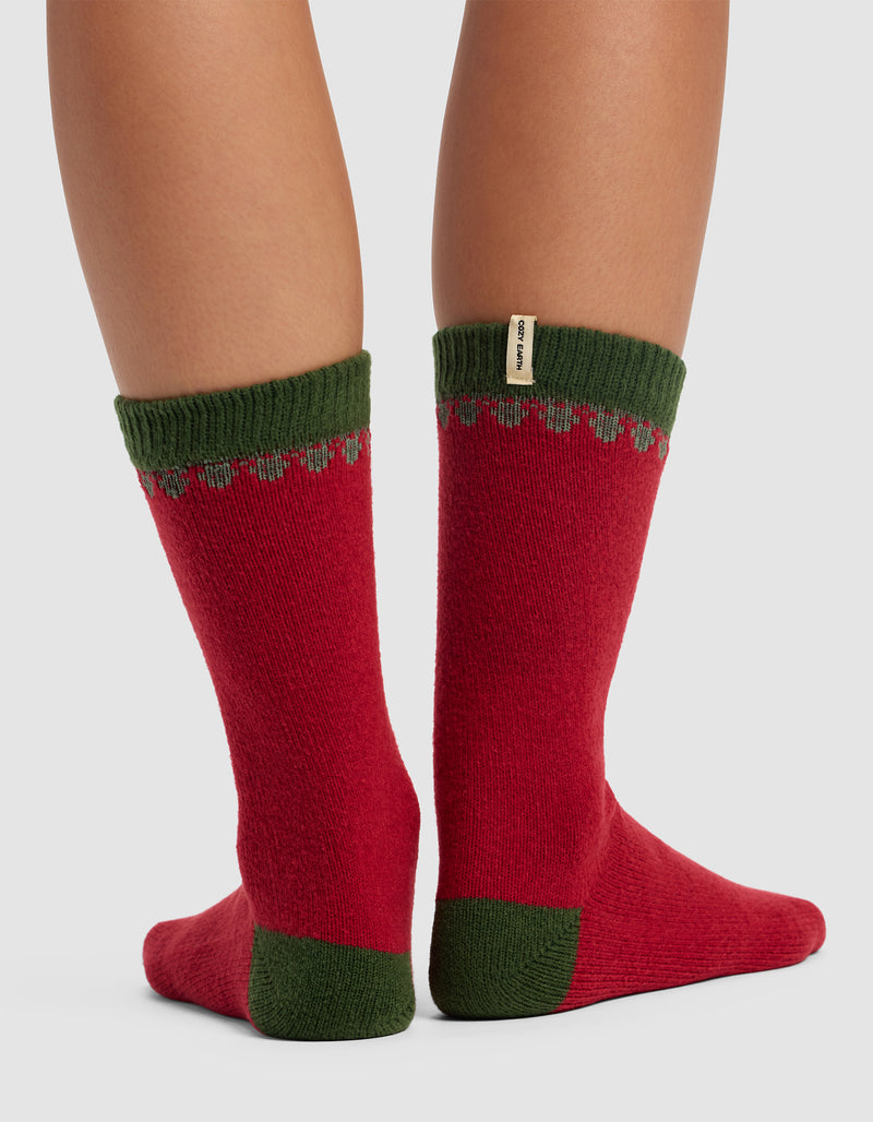 A person modeling Cozy Earth’s The Mistletoe Plush Lounge Sock with red fabric, green cuffs, heels, and toes, a subtle pattern near the top, and a small beige tag on the left sock stands against a neutral background.