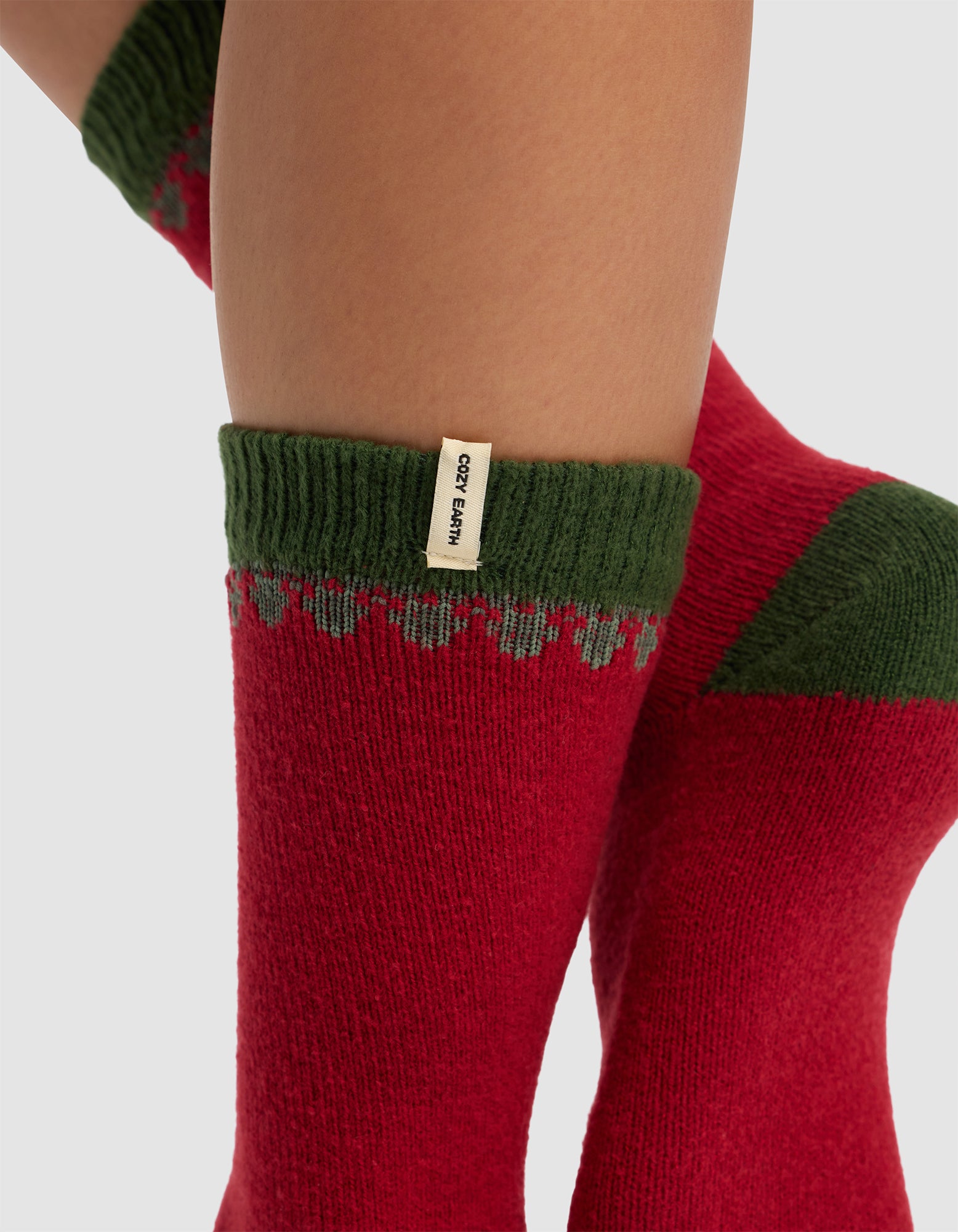 Close-up of a person's legs wearing The Mistletoe Plush Lounge Sock by Cozy Earth—cozy red socks with green cuffs, a small beige label, and a decorative pattern near the top.