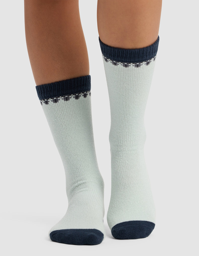 Someone wearing The Mistletoe Plush Lounge Sock by Cozy Earth, featuring light fabric with dark blue toes, heels, and cuffs, plus a decorative blue pattern at the top, stands on a light surface.