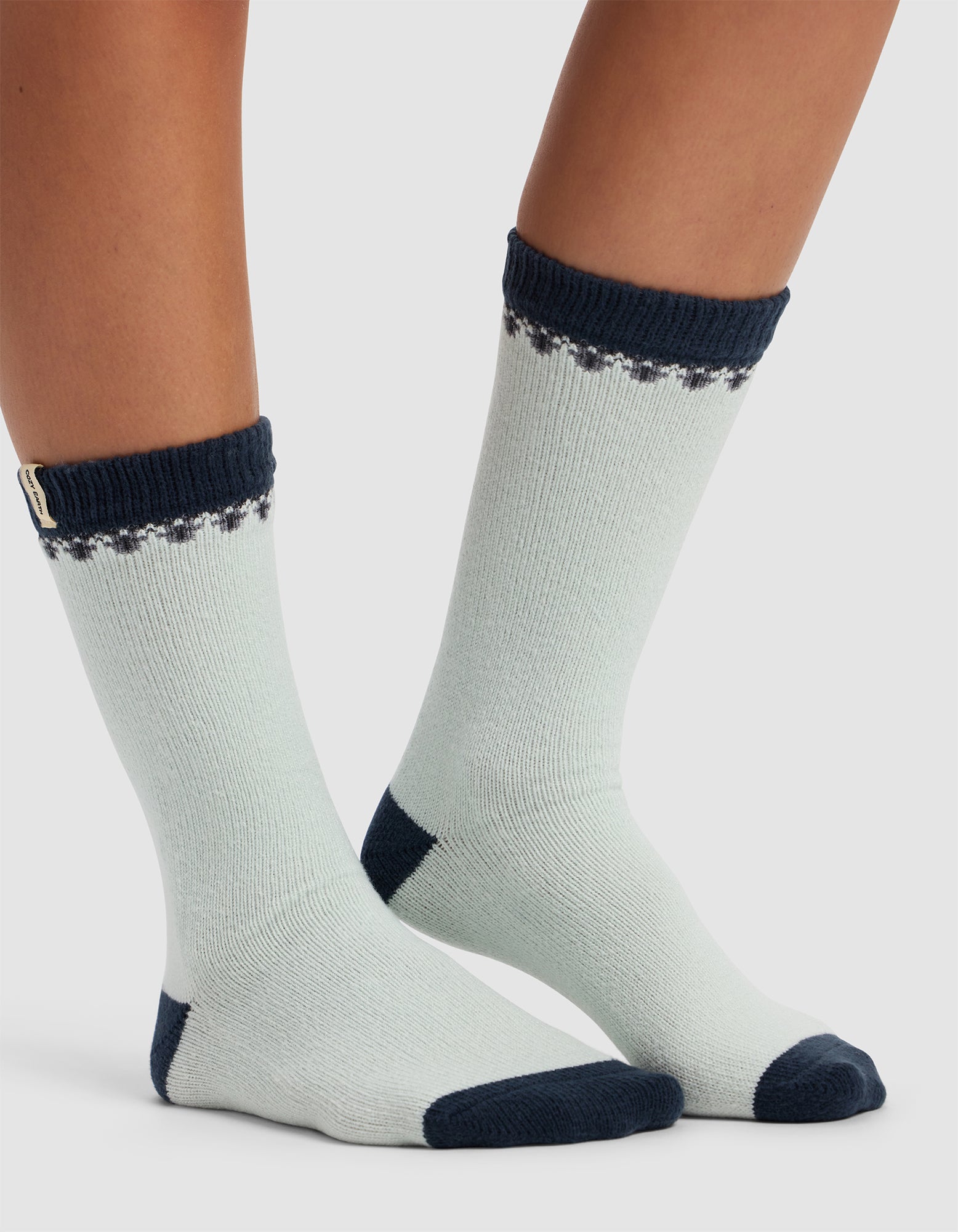 Two feet wearing Cozy Earth's The Mistletoe Plush Lounge Sock in light grey with dark navy-blue cuffs, toes, and heels. The socks feature a small dotted pattern near the top and a branded tag on one cuff. Background is plain white.