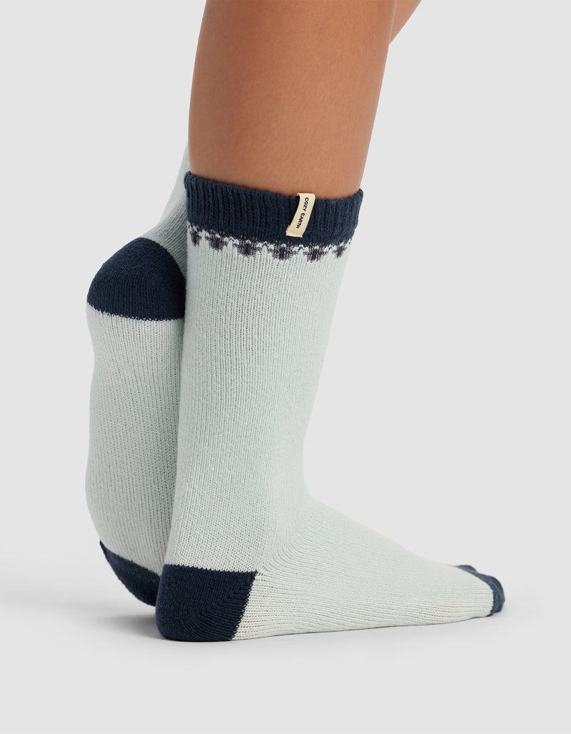 A person wears The Mistletoe Plush Lounge Sock by Cozy Earth—light gray crew socks with navy blue toes, heels, and cuffs, a small decorative pattern near the cuff, and a beige tag. Their feet are crossed at the ankles.